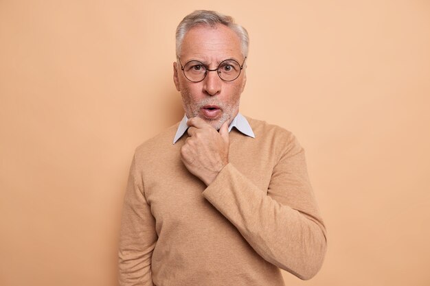 man holds chin reacts emotionally to news looks shocked at camera wears optical round spectacles casual jumper isolated on brown