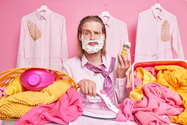 мужчина держит утюг и щетку для бритья занят глажкой одежды для стирки много работает в выходные дни позирует на веревках на розовом