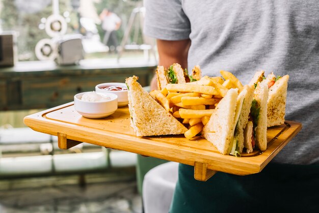 Man holds wooden board club sandwich toast bread chicken tomato cucumber french fries mayonnaise ketchup side view