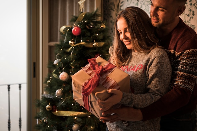 Человек обнимает веселая женщина с подарочной коробке в свитерах возле елки