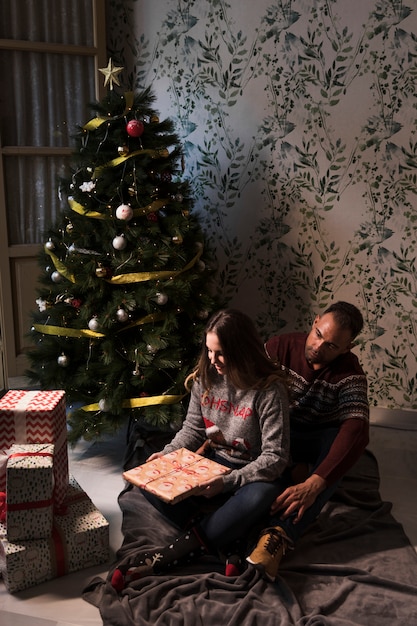 Man hugging woman from back with gift on coverlet near Christmas tree
