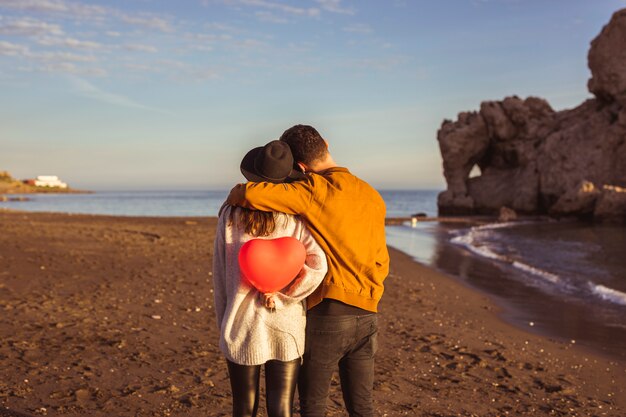 Man hugging woman with red heart balloon on sea shore