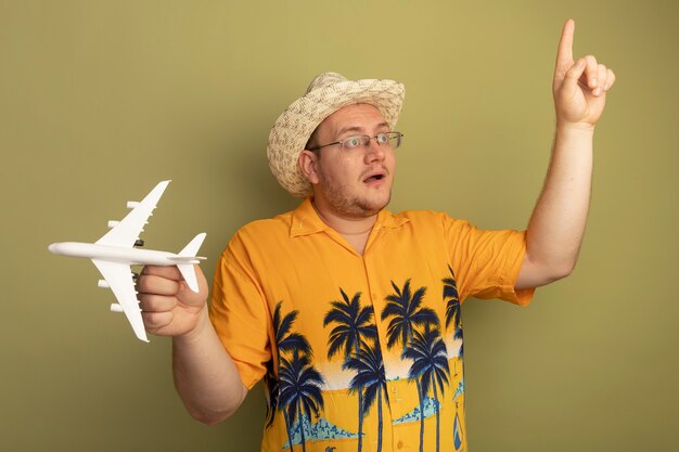 무료 사진 여름 모자에 주황색 셔츠를 입고 안경에 남자가 옆으로 집게 손가락을 보여주는 녹색 벽에 놀란 행복 서를 보여주는 장난감 비행기를 들고