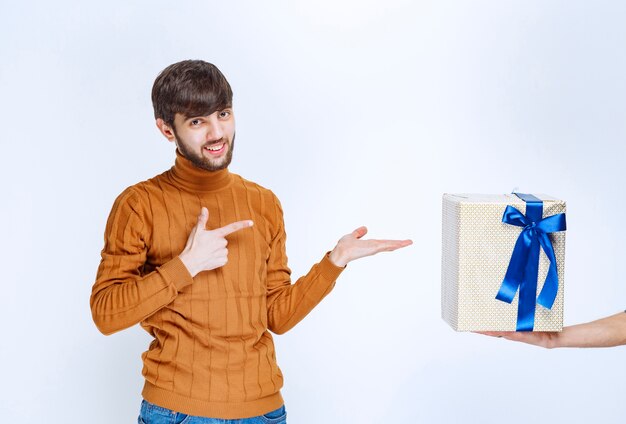 Мужчине предлагают белую подарочную коробку с голубой лентой, и он показывает на нее.