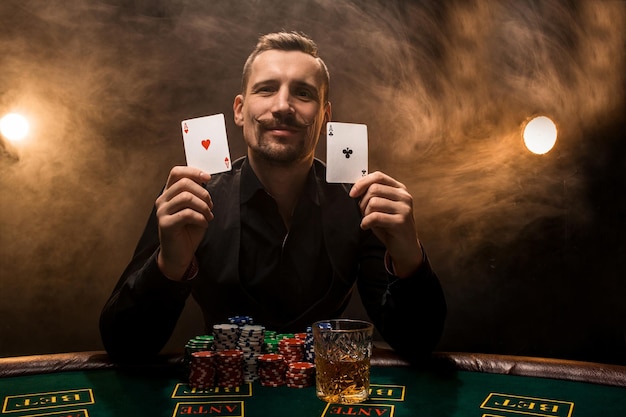 Man is playing poker with a cigar and a whiskey, a man show two cards in the hand, winning all the chips on the table with thick cigarette smoke. The concept of victory