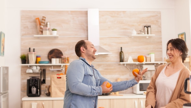 Man juggle with oranges for woman while preparing tasty and nutritious smoothie. Healthy carefree and cheerful lifestyle, eating diet and preparing breakfast in cozy sunny morning