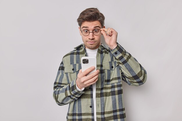 man keeps hand on rim of spectacles stares at smartphone screen reads shocking news has surprised face wears casual checkered shirt isolated on white looks at smething amazing