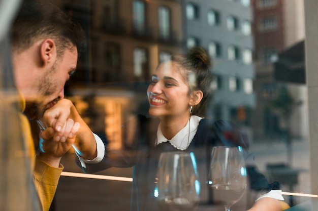 Man kissing hand of smiling woman near glasses of wine in restaurant near window