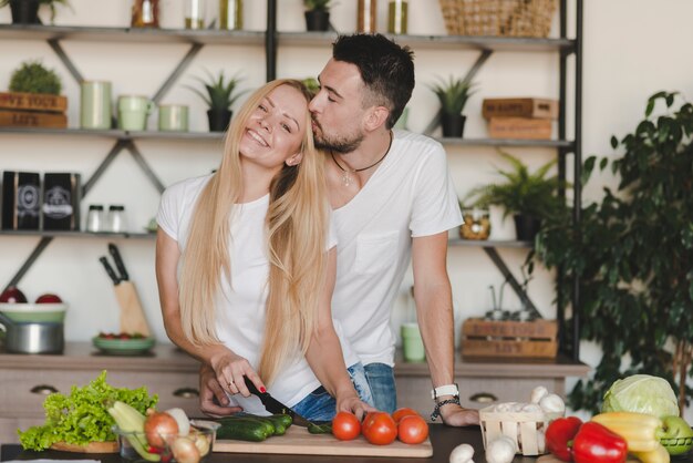 Man kissing her girlfriend cutting vegetables in kitchen