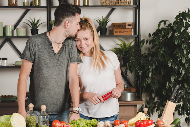 Man kissing her girlfriend on forehead holding red chilies in kitchen