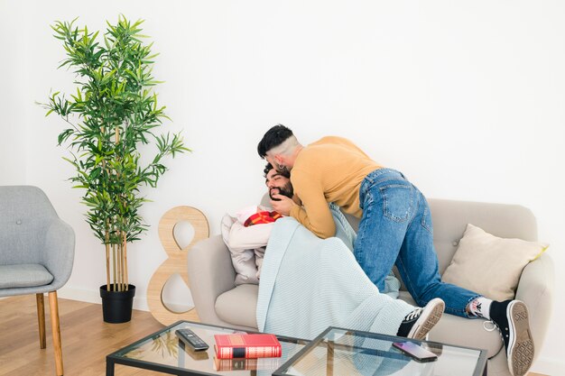 Man kissing his boyfriend lying on sofa with baby in hand at home