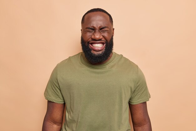  man laughs happily shows white perfect teeth dressed in casual t shirt poses on brown