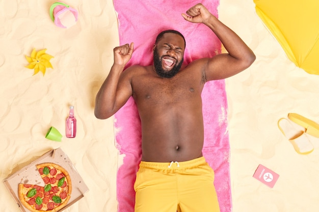  man lies on pink towel makes fist pump poses with bare torso dressed in yellow shorts has fun at beach surrounded by different items celebrates excellent news