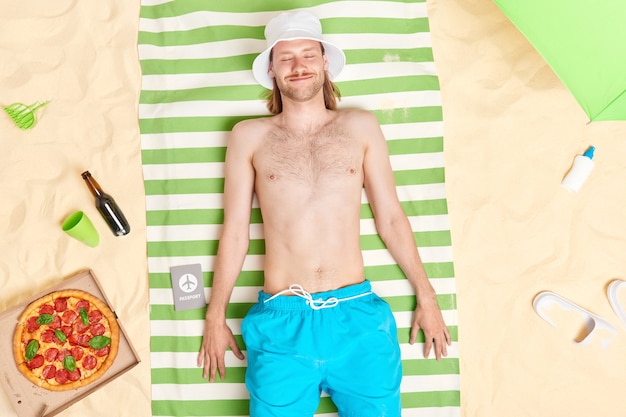  man lies on towel at beach enjoys recreation time poses with closed eyes at seaside surrounded by delicious pizza bottle of bee sunscreen slippers parasol