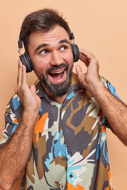 man listens audio track in wireless headphones enjoys perfect sound wears colorful shirt has fun during free time isolated on beige