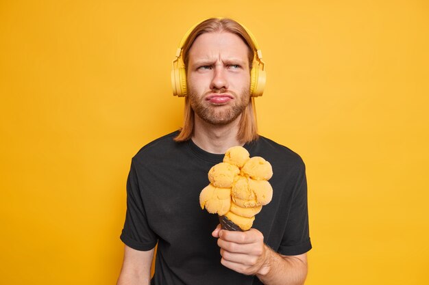 man listens music via stereo headphones holds big cone appetizing ice cream feels displeased dressed in black t shirt isolated on yellow