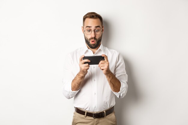 Man looking amazed at mobile phone, standing against white background.