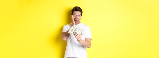 Free photo man looking amazed at money winning cash prize standing against yellow background