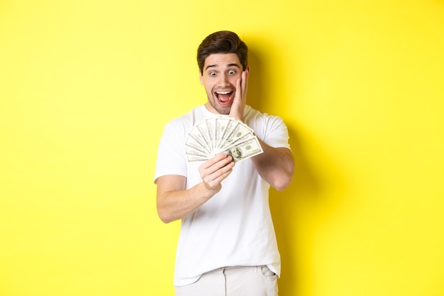 Man looking amazed at money, winning cash prize, standing against yellow background.