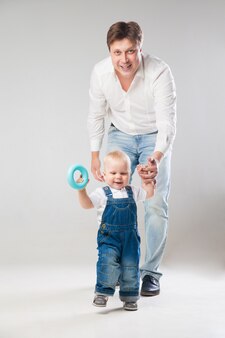 Man looking at camera and holding toddler's hand while baby is making steps on plain grey background.