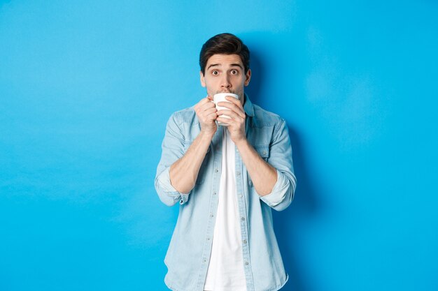 青い背景の上に立って、興奮して白いマグカップからお茶やコーヒーをすすりながら見ている男