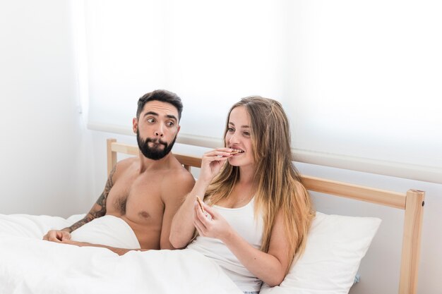 Man looking at his wife eating cookies on bed