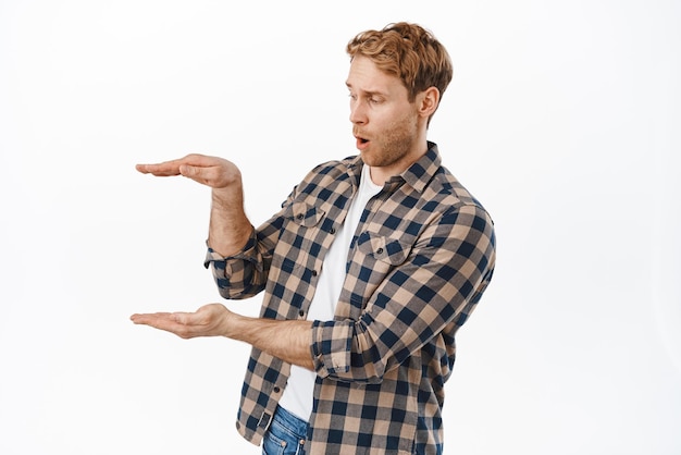 Man looking impressed at object he is holding Redhead guy display an item in hands and look amazed at empty space standing against white background