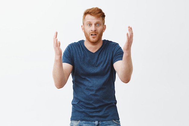 Man looking at what left of his car, feeling shocked, standing over gray wall in stupor. Portrait of stunned disappointed and upset european redhead male, gesturing with palms and dropping jaw