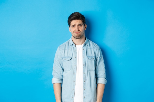 Man looking with doubt at camera, frowning and sulking, feeling uneasy about something, standing against blue background