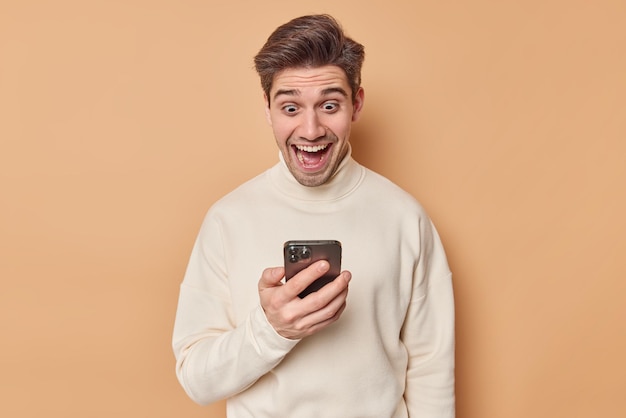 man looks happily at smartphone screen reads amazing notification wears casual turtleneck isolated on beige. Wow what amazing offer. Greatly surprised guy with gadget