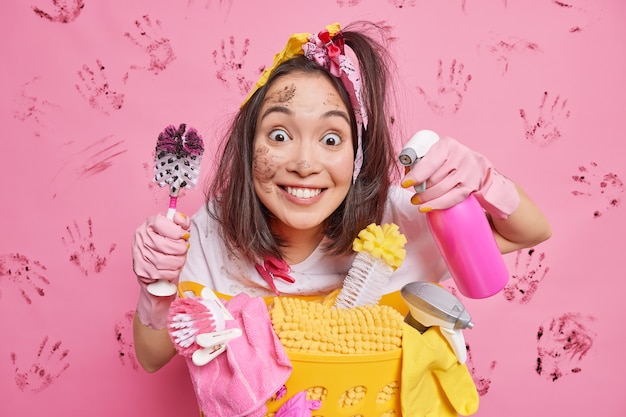 man looks scrupulously smiles positively poses near basket with cleaning supplies holds dirty toilet brush and dispenser bottle isolated on pink