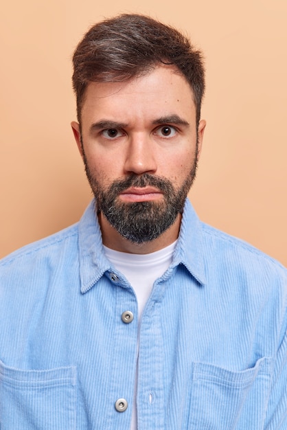 Foto gratuita l'uomo sembra serio ha un'espressione rigorosa guarda direttamente la telecamera indossa una camicia di velluto blu sorride con la faccia isolata sul beige