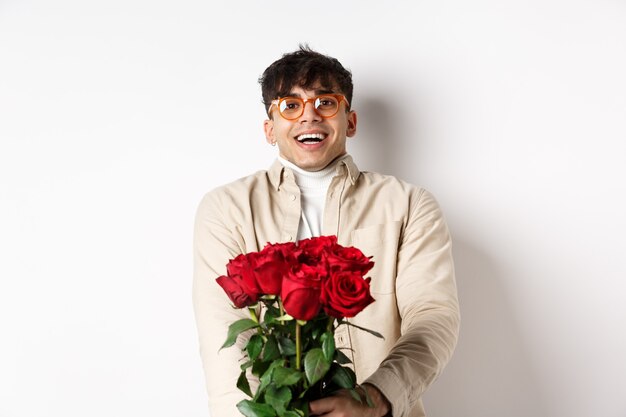 赤いバラを持って、カメラを優しく見て、幸せそうな顔で恋人を見つめ、ガールフレンドとバレンタインデーを祝って、白い背景の上に立っている愛の男