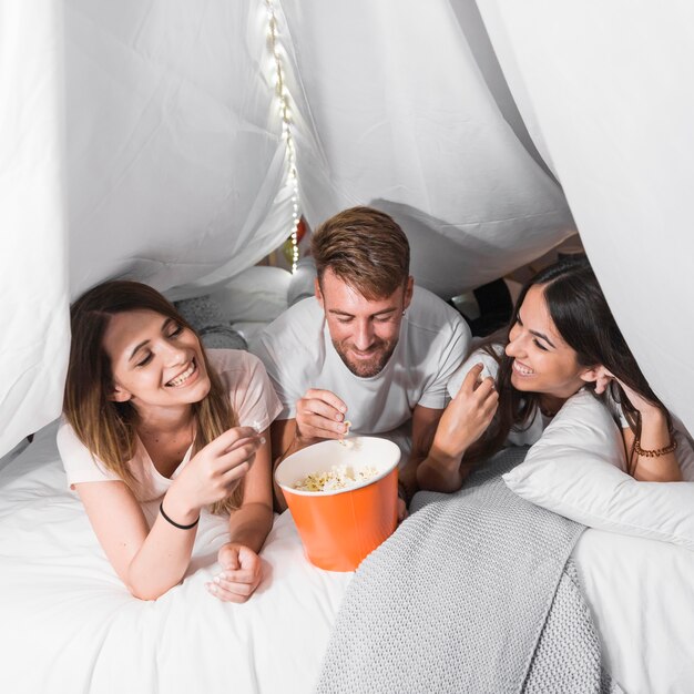 Бесплатное фото Человек, лежащий с двумя подругами на кровати, едят попкорн