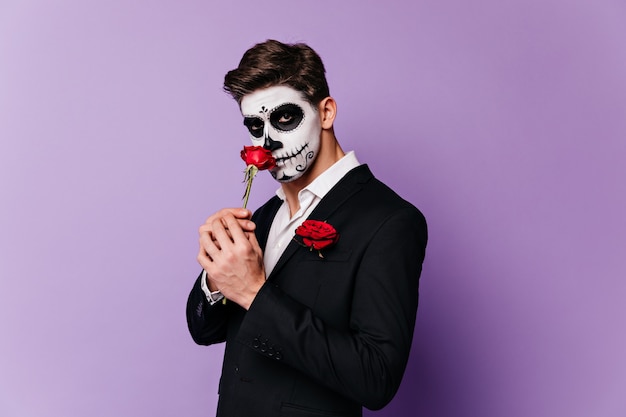 Man in masquerade costume sniffs red big flower and mysteriously looks into camera.