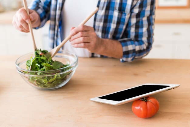 Man mixing the leafy vegetable with wooden tablespoon in the glass bowl with digital tablet and tomato on wooden desk
