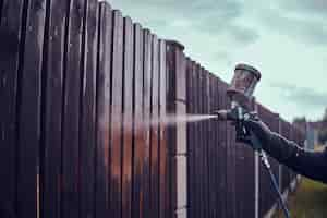 Free photo man n protective gloves is applying paint to the old fence using airbrush at his garden.