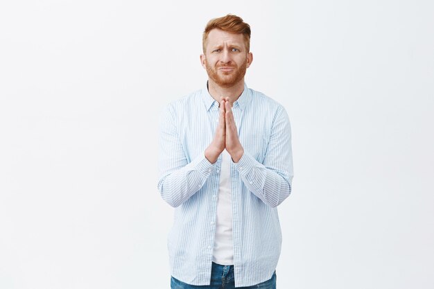 Man in need asking help. Portrait of upset and desperate mature caucasian guy with ginger hair holding hands in pray, frowning and begging for favour or apology, being in trouble over grey wall