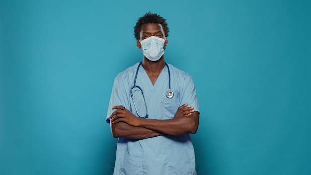 Man nurse standing with crossed arms while wearing face mask for protection against coronavirus pandemic. Medical assistant with healthcare uniform and stethoscope looking at camera.