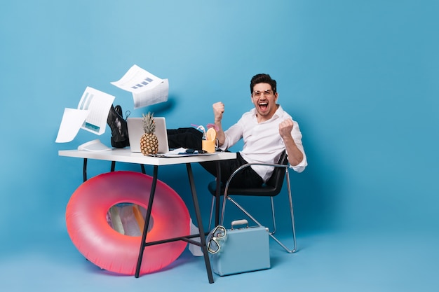 Free photo man in office outfit rejoices while working in laptop amid falling sheets of paper. guy in glasses poses with pineapple, suitcase and mask for diving.