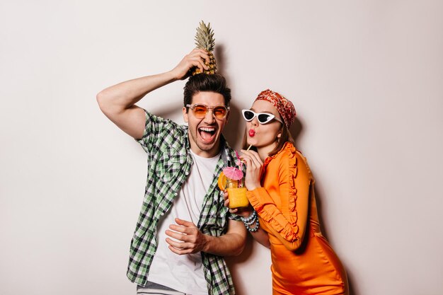 Man in orange glasses holds pineapple on his head and laughs Woman in bright dress and sunglasses is drinking cocktail