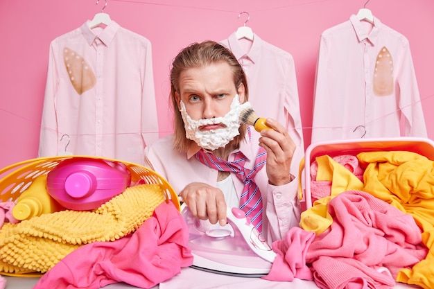 man outs on clothes in morning to work going to business meeting being in hurry shaves and irons clothes simultaneously poses near big piles of laundry