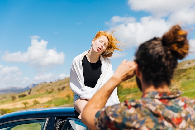 Man photographing grimacing woman sitting on car roof
