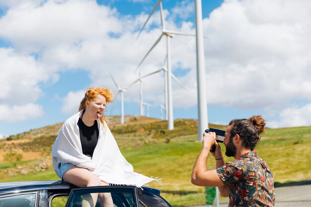 Man photographing grimacing woman in white scarf sitting on car roof