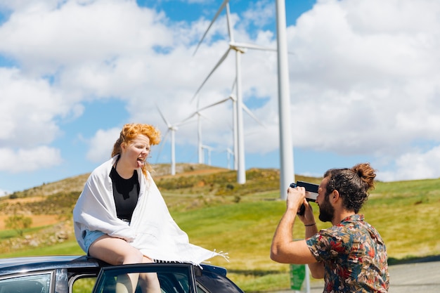 Man photographing grimacing woman in white scarf sitting on car roof