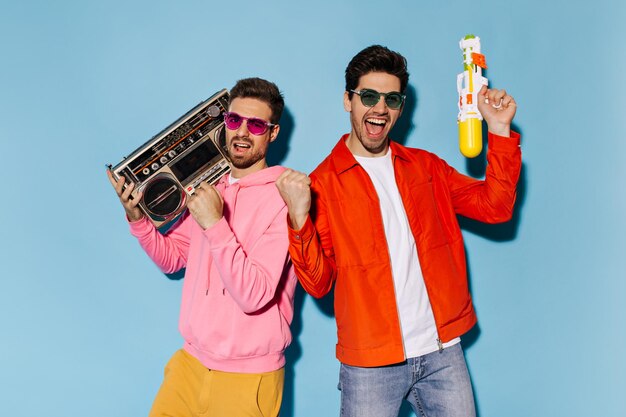 Man in pink hoodie and yellow pants holds record player Cool guy in orange jacket and jeans plays with water gun Friends dance and have fun on blue background