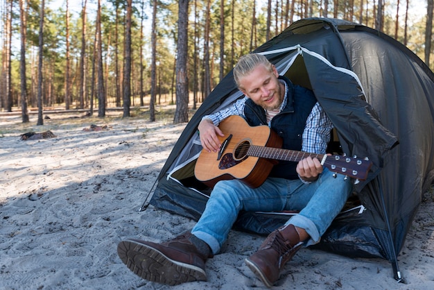 Человек играет на акустической гитаре и сидит в своей палатке