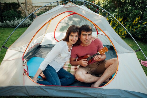 Man playing ukulele sitting with his wife in tent looking at camera