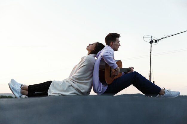 Man plays a guitar while his woman leans to him tender on the rooftop