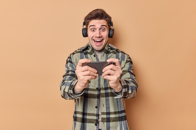 男はモバイルビデオゲームをプレイし、スマートフォンを水平に保持し、ワイヤレスヘッドフォンを着用し、ベージュで隔離された市松模様のシャツを着用します。人と技術の概念